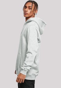 Gray hoodie made of soft fabric, features a large hood, front pocket, and ribbed cuffs. Model stands sideways against a neutral background.