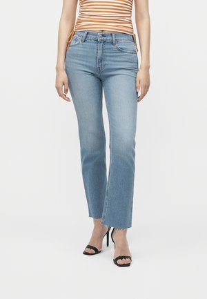 Femme portant un jean droit bleu clair à bords francs, des talons noirs ouverts et un haut à rayures orange et blanc sur fond blanc.