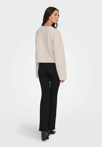 Przycięta, jasno beżowa bluzka z szerokimi rękawami; zestawiona z czarnymi spodniami dzwonami i czarnymi botkami na obcasie. Widok z tyłu, jednolite tło.
