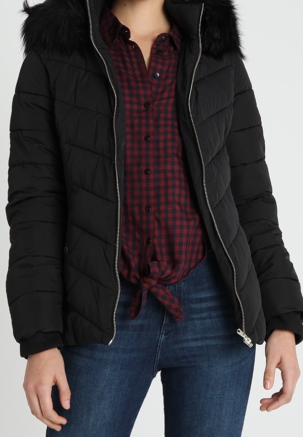 Svart puffjacka med huva av fuskpäls, blixtlås, och quiltat mönster, bärs över en röd och svart rutig skjorta, ihop med blå jeans.