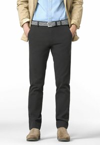 Mann trägt ein hellblaues Hemd, eine beige Jacke, einen gemusterten Gürtel, dunkelgraue Hosen und tanfarbene Wildlederschuhe und steht mit den Händen in den Taschen.