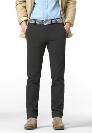 Mann trägt ein hellblaues Hemd, eine beige Jacke, einen gemusterten Gürtel, dunkelgraue Hosen und tanfarbene Wildlederschuhe und steht mit den Händen in den Taschen.