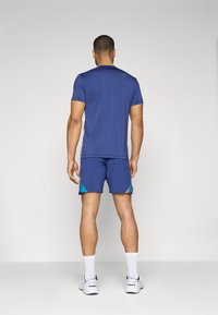 Sportliches Männeroutfit mit einem blauen T-Shirt mit kurzen Ärmeln und passenden Shorts mit türkisfarbenen Akzenten, kombiniert mit weißen Socken und Turnschuhen.