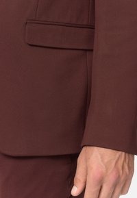 Bordeauxfarbener Anzugjacke im Close-up, mit glatter Textur, strukturiertem Design und einer einzelnen Brusttasche, die die maßgeschneiderte Passform und klaren Linien betont.