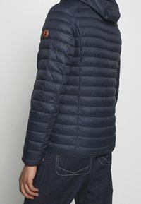 Giacca puffer blu navy con design trapuntato testurizzato e cappuccio. Presenta un'etichetta con logo arancione sulla manica sinistra e una silhouette aderente.