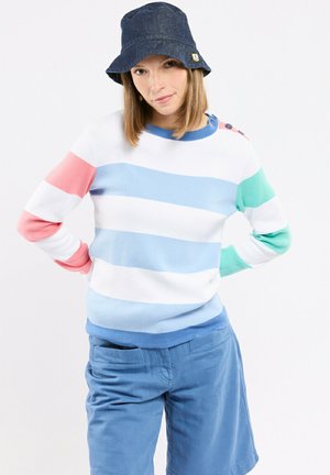 Femme portant un chapeau bob en jean bleu, un pull blanc avec des rayures pastel rose, bleu et vert, et un pantalon bleu clair, les mains sur les hanches.