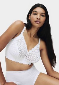 Bílá krajková braletka s květinovou výšivkou, vroubkovanými okraji a nastavitelnými ramínky. Nošená s odpovídajícím bílým spodním prádlem.