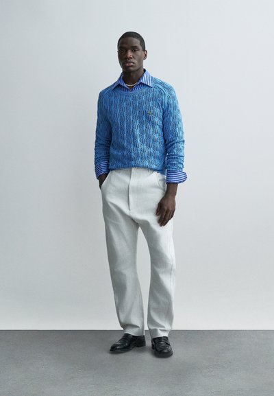 Homme debout portant un pull texturé bleu clair par-dessus une chemise à rayures bleues, un pantalon gris clair, des chaussures noires et un collier de perles.