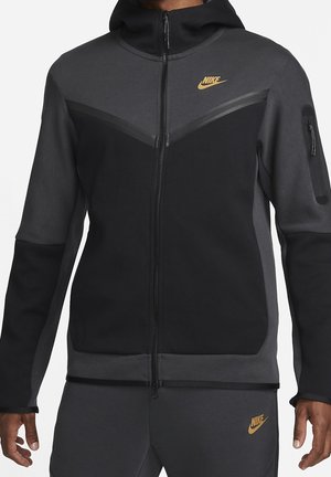 Mężczyzna w czarnej i ciemnoszarej bluzie Nike na zamek oraz pasujących spodniach z złotymi logo Nike na piersi i udzie.