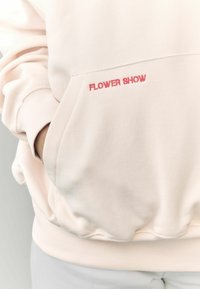 Šviesiai rožinė džemperis su raštuotomis rankogaliais ir apačia. Yra priekinė kišenė ir raudonai siuvinėtas "FLOWER SHOW" užrašas ant kišenės.
