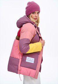 Barevná péřová bunda s panely růžové, fialové a žluté barvy. Má kapuci, druky a viditelnou etiketu na přední kapse.
