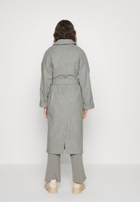 Vrouw met lang bruin haar, gekleed in een grijze wollen jas met riem, grijze broek en beige chunky sneakers, staand tegen een eenvoudige witte achtergrond.