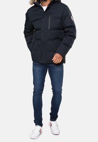 Navyblå pufferjakke med hætte beklædt med pels, der har knapper foran, en lynlåslomme og quiltet tekstur. Kombineret med blå jeans og hvide sneakers.