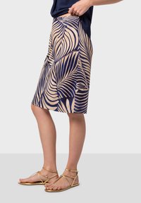 Falda azul marina con patrón de hojas de palmera beige, de longitud media, hecha de tela ligera. Combinada con sandalias doradas de tiras.
