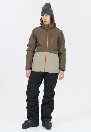 Vanntæt jakke i brun og beige, med en orange lynlås, hætte og justerbare manchetter. Sat sammen med sorte skibukser og støvler.