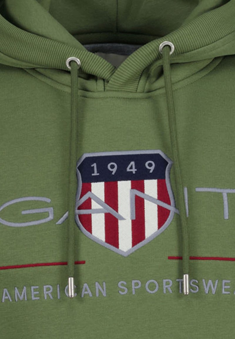 Felpa verde con cappuccio e coulisse. Presenta un logo a scudo con strisce rosse e bianche e la scritta "GANT" in grigio.