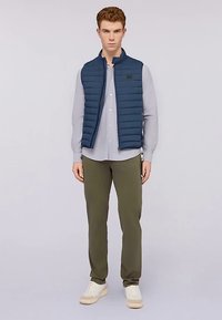 Námořnická prošívaná vesta přes světle šedou košili na knoflíky, doplněná olivově zelenými kalhotami a bílými teniskami. Hladká textura a minimalistický design.