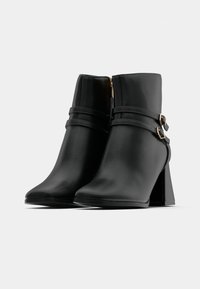 Mustad mustad jalanõud, mis on valmistatud siledast sünteetilisest materjalist, millel on terav nina, plokknina ja dekoratiivsed rihmad, millel on kuldsed riistad.