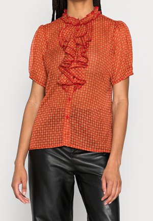 Blusa rossa con motivo, dotata di maniche corte, colletto arricciato e chiusura con bottoni. Realizzata in un tessuto leggero e trasparente. Abbinata a pantaloni neri.