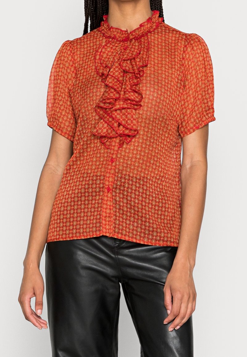 Blusa rossa con motivo, dotata di maniche corte, colletto arricciato e chiusura con bottoni. Realizzata in un tessuto leggero e trasparente. Abbinata a pantaloni neri.