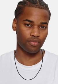 Zwarte ketting gedragen over een wit T-shirt. Vloeiende metalen textuur, minimalistisch ontwerp met een eenvoudige sluiting. Subtiele glans.