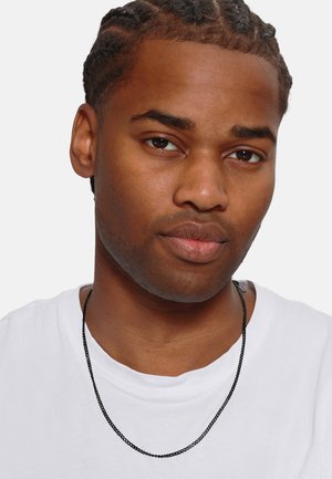 Schwarze Halskette aus Kette, getragen über einem weißen T-Shirt. Glatte metallische Textur, minimalistisches Design mit einem einfachen Verschluss. Subtile Ausstrahlung.