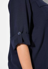 Camisa azul marino con mangas remangadas sujetas por un botón. Hecha de un material texturizado con un corte relajado, presenta un detalle de bolsillo en el pecho.