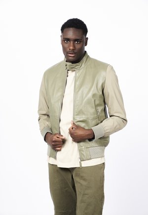 Chaqueta de cuero verde oliva clara con dobladillo y puños acanalados, que presenta un cuello alto y bolsillos con cremallera, superpuesta sobre una camisa beige.