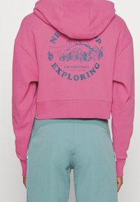 Felpa cropped rosa con cappuccio, caratterizzata da una grafica blu sulla schiena e polsini a costine. Indossata sopra pantaloni azzurri chiari con zip.