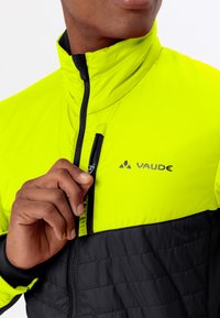 La mano di un uomo che chiude la cerniera di una giacca a collo alto gialla e nera con il logo Vaude sul lato sinistro del petto.