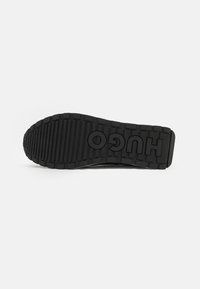 Semelle de chaussure en caoutchouc noir avec des rainures profondes et un logo "HUGO" bien visible. Surface texturée pour une meilleure adhérence, présentant un motif défini pour la traction.