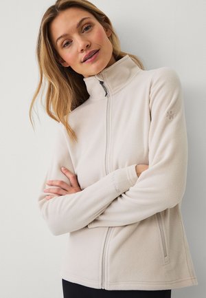 Kobieta o blond włosach, ubrana w beżową polarową kurtkę z wysokim kołnierzem i zamkiem błyskawicznym z przodu, ze skrzyżowanymi ramionami, na jednolitym jasnym tle.