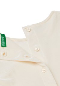 Zbliżenie na dekolt kremowego aksamitnego ubrania z trzema perłowymi guzikami i zieloną etykietą United Colors of Benetton.