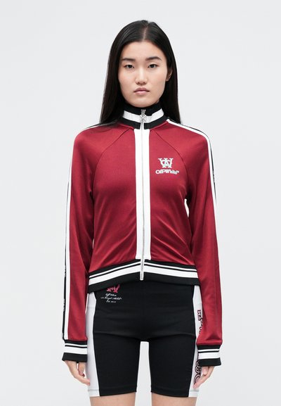 Veste de survêtement rouge avec accents blancs, fermeture éclair à l'avant, col montant et logo sur la poitrine. Associée à un short noir avec détail graphique.