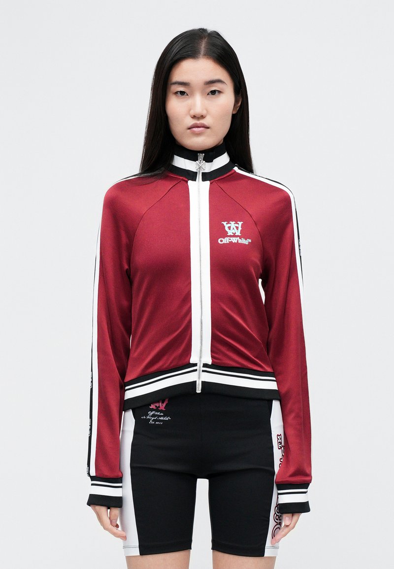 Giacca da allenamento rossa con dettagli bianchi, zip frontale, collo alto e logo sul petto. Abbinata a pantaloncini neri con dettagli grafici.