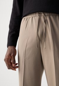 Pantalons beiges légers avec une taille élastique et un cordon de serrage. Texture lisse et coupe décontractée, associés à un pull noir.