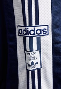 Dres Adidas s navy modrou sieťovinou, bielymi bočnými pruhmi a vyšívanými logami: "adidas" v modrej farbe a logo Trefoil s textom.