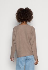 Person mit lockigem Haar, der einen beigen langärmligen Rippstrickpullover und hellblaue Jeans trägt und vor einer schlichten weißen Wand steht.