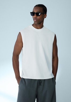 Mand iført hvid ærmeløs skjorte, mørke solbriller, mørke shorts med hænderne i lommerne, stående mod en lysegrå baggrund.