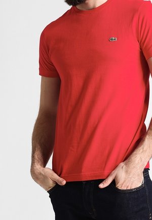 Homme portant un t-shirt rouge à manches courtes avec un petit logo crocodile et un jean bleu foncé, mains dans les poches devant un fond uni.