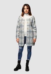 Abrigo largo de cuadros en tonos grises con acentos blancos y negros, combinado con un suéter de cuello alto blanco y unos vaqueros azul desgastados, terminado con botas negras hasta el tobillo.