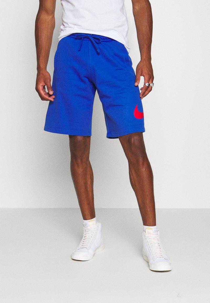 Blå bomullsshorts med elastisk midjeband och snörning. Har en stor röd Nike-logga på vänster sida. Bärs med vita sneakers.