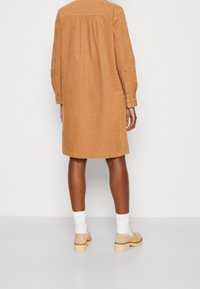 Langärmliges, hellbraunes Cordhemdkleid mit gerafftem Rückendetail und geknöpften Manschetten, getragen mit hellen Socken und beigen, klobigen Schuhen mit Absatz.