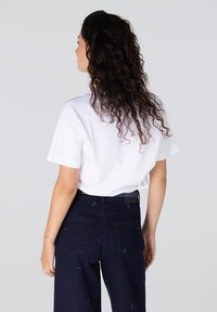 Witte korte mouwen t-shirt gecombineerd met donkerblauwe wijde jeans met kleine blauwe patronen en zakken, gezien vanuit de achterkant.