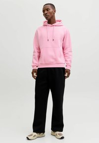 Felpa con cappuccio rosa in tessuto morbido, con tasca a marsupio e coulisse regolabile. Abbinata a pantaloni della tuta neri e sneaker beige.