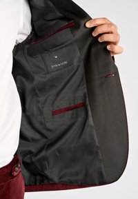 Hand houdt een open donker kostuumjasje vast, waarbij de zwarte satijnen voering met bordeauxrode bies en het label "JUNK de LUXE" op het binnenzakje zichtbaar zijn.