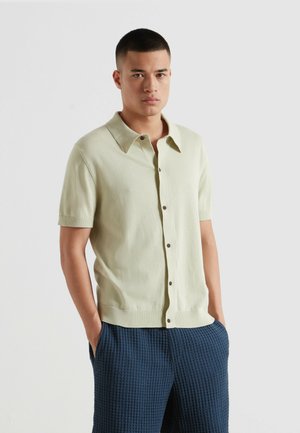 Mann mit kurzem Haar, der ein beigefarbenes Kurzarm-Hemd mit Knöpfen und strukturierte marineblaue Hose trägt, Hände in den Taschen, vor einfachem hellem Hintergrund.