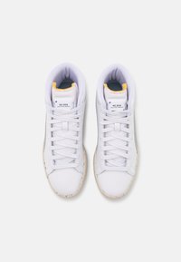 Paire de baskets montantes blanches avec des lacets blancs, des semelles tachetées blanc cassé et une doublure jaune à l'ouverture de la chaussure, vue de dessus.