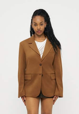 Γυναίκα με μακριά πλεξούδα που φορεί καφέ υπερμεγέθη σακάκι πάνω από λευκό δαντελένιο μπλουζάκι και σκοτεινή φούστα, στέκεται μπροστά σε απλό φόντο.