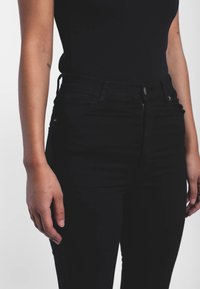 Černé džíny s vysokým pasem, hladkým povrchem, se předními kapsami, zipovým zapínáním a přiléhavým střihem, vystavené na neutrálním pozadí.